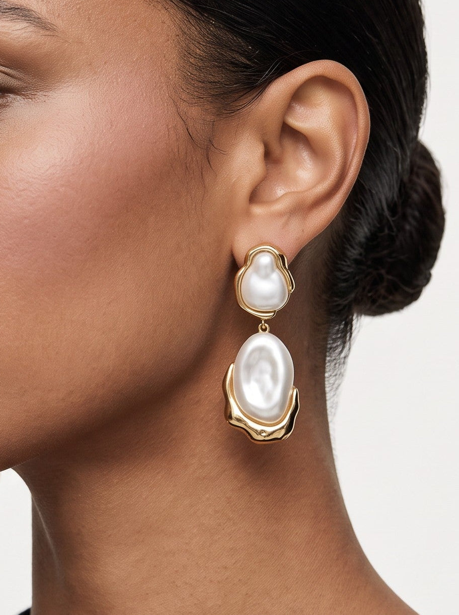 A woman in profile wearing a pair of gold drop earrings with large white pearl-like oval charms, shown against a light neutral background.